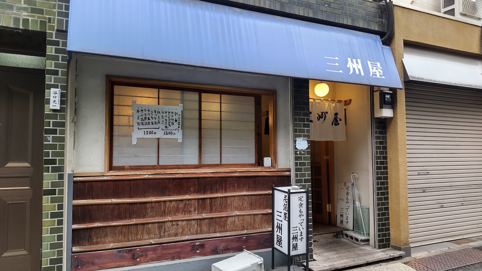 青い日よけに白く三州屋と書かれた店先。木の格子窓と暖簾、入口の行灯には居酒屋 三州屋、定食もやっていますの文字。