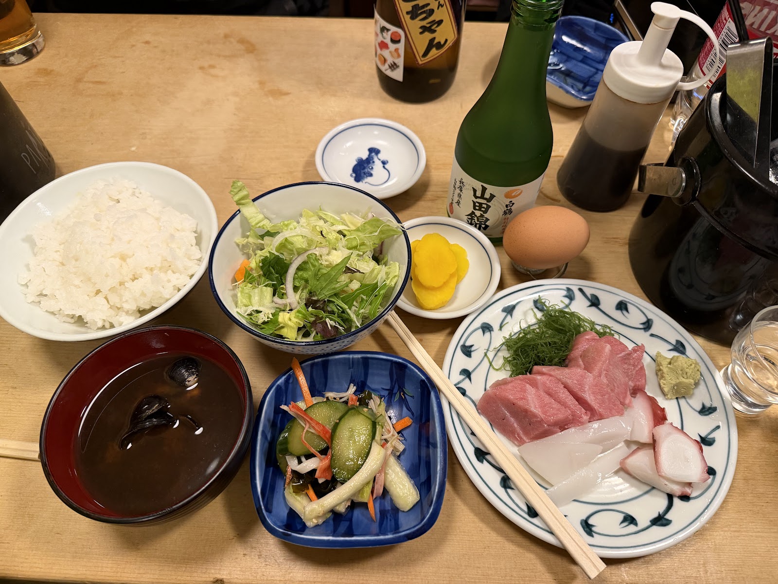 木のカウンターに並んだ定食。ご飯と味噌汁、サラダ、漬物、皿に盛った刺身。奥に瓶と卵。