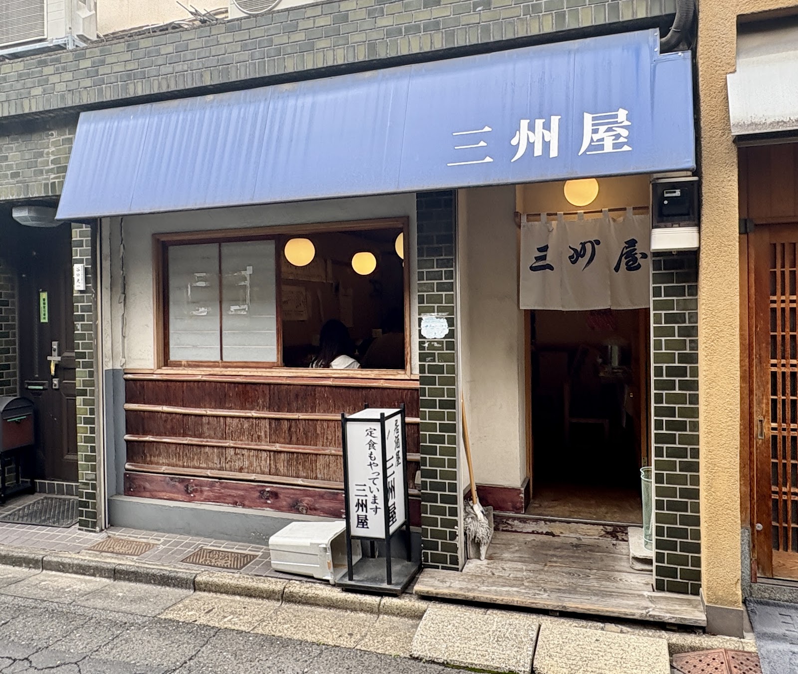 通りから見た店先。青い日よけと暖簾に三州屋、行灯、木の格子窓、緑のタイルの柱。